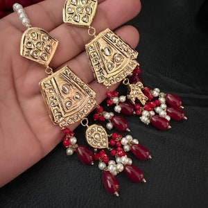 May include: Gold earrings with red and white beads. The earrings feature a large, ornate, gold pendant with intricate details and a smaller, teardrop-shaped pendant.