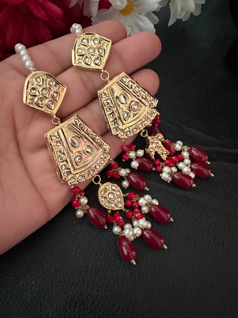 May include: Gold earrings with red and white beads. The earrings feature a large, ornate, gold pendant with intricate details and a smaller, teardrop-shaped pendant.
