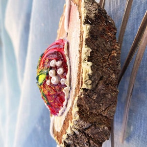 Könnte beinhalten: Ein roter und grüner bemalter Holzschmuck mit weißen Perlen und einem weißen Quasten, der an einer natürlichen Holzscheibe hängt.