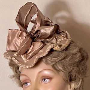 May include: A vintage-style hat in shades of tan and brown, featuring a large bow and decorative trim. The hat is made of a satin-like material and is likely from an earlier era. The design includes a decorative trim around the base of the hat.