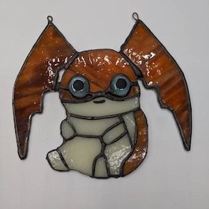 May include: Stained glass art piece depicting a cartoon-like creature with large, orange wings and blue eyes. The creature's body is primarily off-white, with orange accents. The piece is framed with dark metal and has two loops for hanging.