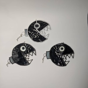 Może przedstawiać: Trzy czarne i białe ozdoby ze szkła witrażowego w kształcie postaci z gry wideo z dużymi ustami i jednym okiem. Ozdoby wiszą na srebrnych łańcuszkach.