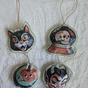 May include: Four hanging ornaments featuring vintage-style illustrations: a black cat face, a skull inside a crystal ball, a crescent moon with a pumpkin, and a devil face. Each ornament is made of fabric with a green stitched edge and a jute string.