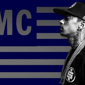 May include: A black and white photo of a person wearing a black baseball cap with an American flag patch, a black shirt, and a silver chain necklace. The person is looking to the right. The background is a blue and gray striped pattern with the text "TMC" in gray.