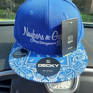May include: A blue and white baseball cap with a paisley bandana pattern on the brim. The front of the cap has white embroidery that reads "Naybors & Gang Stay Dangerous 100". The back of the cap has a black and white label that reads "DECKY SNAPBACK".