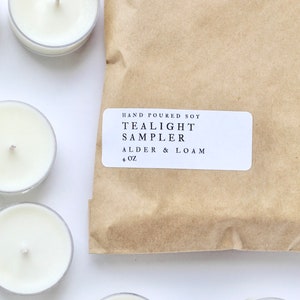 Puede incluir: Un conjunto de velas de té blancas en vasos de plástico transparente, dispuestas alrededor de un paquete de papel marrón. El paquete tiene una etiqueta blanca que dice: "Hand Poured Soy Tealight Sampler Alder & Loam + 4 oz."