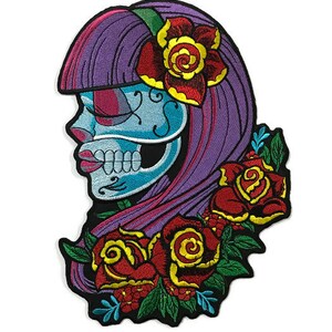 May include: Embroidered iron-on patch featuring a blue and white skull with pink and purple hair, wearing a red and yellow flower. The skull is surrounded by red and yellow roses with green leaves.