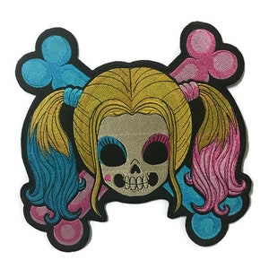May include: Embroidered patch featuring a skull with pigtails, wearing a pink and blue outfit. The skull has a pink heart on its cheek and is surrounded by two crossed bones.