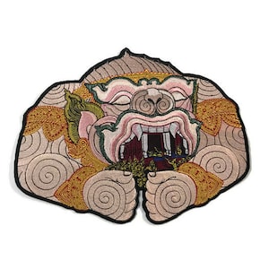 Könnte beinhalten: Ein gestickter Patch, der eine detaillierte Darstellung einer mythischen Kreatur mit einem grimmigen Ausdruck zeigt. Die Kreatur hat ein rosafarbenes Gesicht mit einem großen, offenen Maul, das scharfe Zähne enthüllt. Der Patch ist mit komplizierten Mustern in Gold, Rot und Blau verziert.