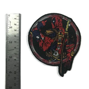 May include: Embroidered patch featuring Deadpool in a circular design. The patch is primarily red and black, with details of the character's costume and a sword. The patch measures approximately 7.6 cm in height.