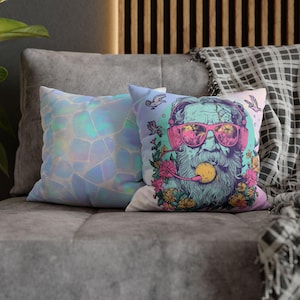 May include: Two decorative throw pillows on a gray couch. One pillow features an abstract geometric pattern in shades of blue and white. The other pillow displays a colorful illustration of a bearded figure wearing sunglasses and headphones, surrounded by flowers and birds.