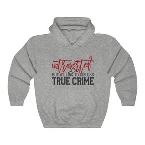 Puede incluir: Una sudadera con capucha gris con el texto "introvertido pero dispuesto a discutir crímenes reales" en rojo y negro. El diseño incluye una lupa. La sudadera es de estilo casual.