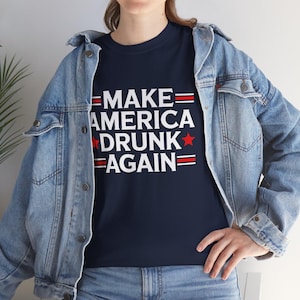 Könnte beinhalten: Marineblaues T-Shirt mit dem weißen Schriftzug "MAKE AMERICA DRUNK AGAIN", akzentuiert mit roten Streifen und einem roten Stern. Das Shirt wird mit einer hellblauen Jeansjacke und einer blauen Jeans getragen.