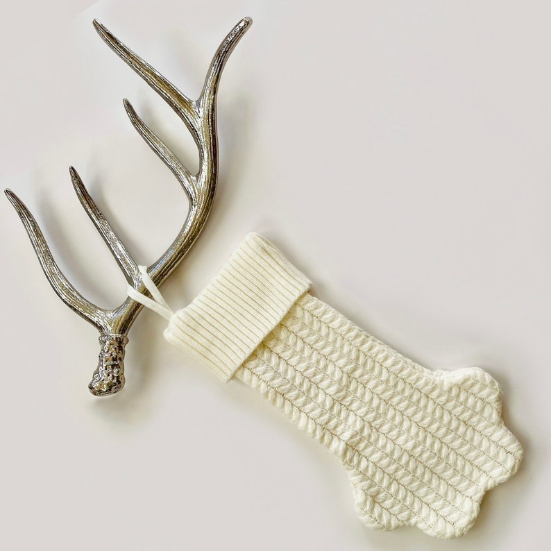 Op de afbeelding: Een witte gebreide kerstsok met een kabelbreipatroon hangt aan een zilveren hertengeweihaak.