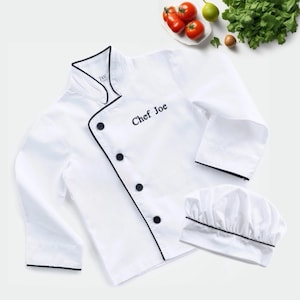 May include: A white chef's jacket and hat set. The jacket has black trim, black buttons, and the embroidered name "Chef Joe". The chef's hat also has black trim. The set is displayed with tomatoes and herbs.