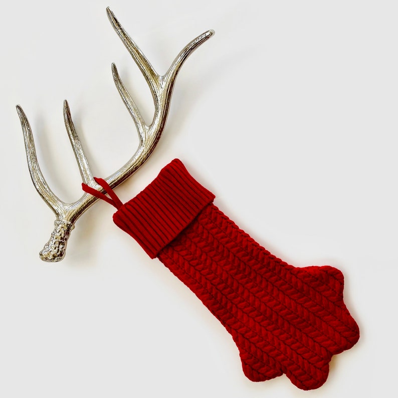 Op de afbeelding: Een rode gebreide sok met een kabelbreipatroon en een boord. De sok is gevormd als een poot en heeft een lus om op te hangen.