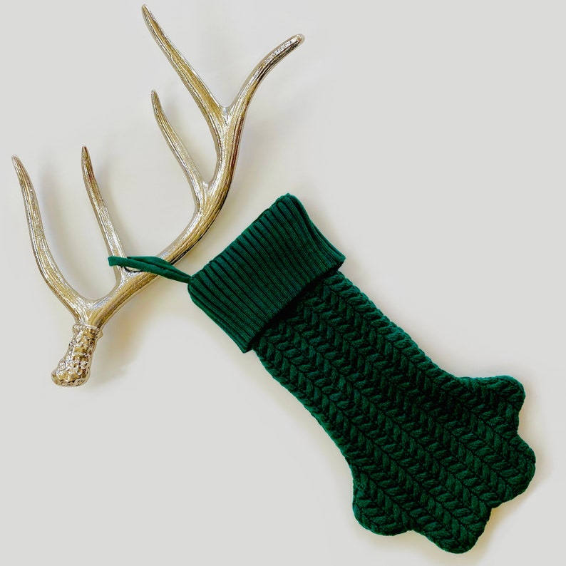 Op de afbeelding: Een groene gebreide kous met een puntige teen en een manchet. De kous hangt aan een zilveren geweistuk.