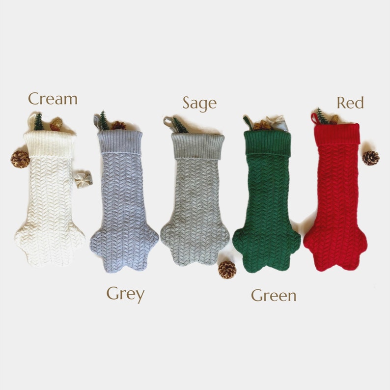 Op de afbeelding: Vijf gebreide kerstsokken in verschillende kleuren: cr&egrave;me, grijs, salie, groen en rood. De sokken hebben een kabelbreipatroon en een puntige teen.
