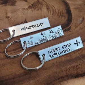 May include: Three silver keychains with engraved designs. The first keychain says "Wanderlust" and has a small tree design. The second keychain has a mountain scene with a bear and trees. The third keychain says "Never Stop Exploring" and has a compass design.