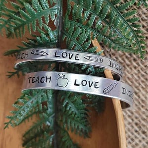 May include: Two silver bracelets with engraved text. The top bracelet reads "Love, Inspire, Teach" and the bottom bracelet reads "Teach, Love, Inspire". Both bracelets feature a pencil and apple icon.