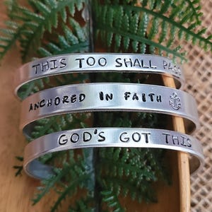 May include: Three silver metal bracelets with inspirational messages. The top bracelet reads "This too shall pass", the middle bracelet reads "Anchored in faith" with an anchor symbol, and the bottom bracelet reads "God's got this".