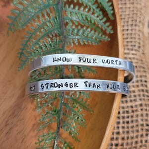Op de afbeelding: Twee zilveren metalen armbanden met inspirerende boodschappen. De bovenste armband heeft de tekst "Know Your Worth" en de onderste armband heeft de tekst "Be Stronger Than Your Excuses".