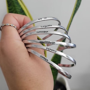 May include: A collection of silver-toned metal bangles, each inscribed with an inspirational word: "let go," "balance," "breathe," "enough," "strong," and "dream." The bangles are held in a hand, with a ring visible on one finger. A green plant is in the background.