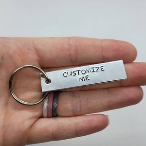 Könnte beinhalten: Silberner Schlüsselanhänger mit einem rechteckigen Anhänger, auf dem "CUSTOMIZE ME" steht.