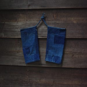 May include: A pair of dark blue denim arm sleeves featuring visible patchwork construction with varying shades of indigo fabric. The two sleeves are connected at the top by a thin denim cord, allowing them to be hung together as a single unit.