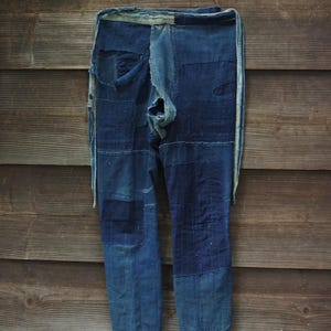 Puede incluir: Un pantalón teñido de índigo con costuras estilo sashiko, múltiples parches en varios tonos de azul, bordes deshilachados y una cinturilla de tela clara con largos lazos laterales que cuelgan desde la línea de la cintura.
