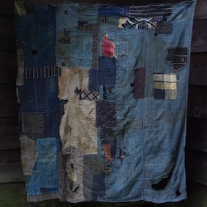 Puede incluir: Un textil rectangular compuesto por varios parches de tela en tonos azules cosidos entre sí. La pieza presenta una mezcla de cuadrados de algodón índigo lisos, a rayas y estampados con remiendos visibles, bordes deshilachados y pequeños agujeros.