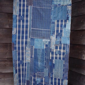 以下が含まれることがあります： 青と白の色合いのパッチワーク織物で、ストライプやチェックなどの様々な模様が特徴です。生地は、手作りの質感を作り出すために縫い合わされた、多数の長方形のパッチで構成されています。木製の背景に吊るされています。