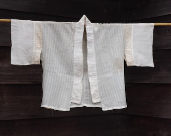 May include: A white, open-front kimono-style jacket with short sleeves, hanging on a wooden rod. The jacket has a simple design with vertical stripes and a collar. The background is a dark wooden wall.