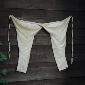 May include: Cream-colored antique pants with a high waist and long legs, hanging against a dark wooden background. The pants have a simple design with seams and ties at the waist.
