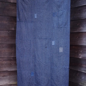 Op de afbeelding: Een indigo blauw textiel met verticale strepen en zichtbare patches van verschillende groottes. Het textiel, mogelijk een quilt of wandkleed, is opgehangen aan een bamboestok. De achtergrond is een houten muur.