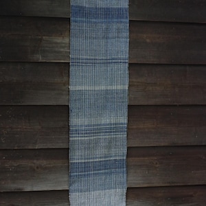 Puede incluir: Un camino de mesa rectangular de tejido trenzado con un patrón de rayas verticales en varios tonos de azul y blanco roto. La tela tiene una apariencia texturizada con variaciones de hilo visibles y un borde deshilachado con flecos.