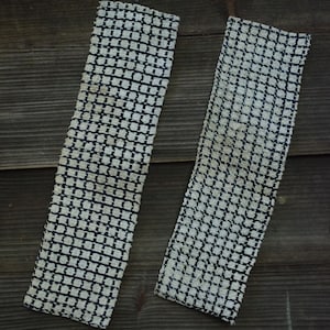 May include: Two rectangular fabric strips featuring a woven grid pattern in black and off-white. The textile has a textured, coarse appearance with a repeating square design throughout the length of both pieces, which are positioned vertically.