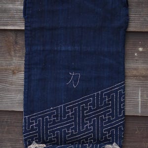 Peut inclure: Sac textile bleu indigo foncé avec des motifs géométriques cousus en blanc le long du bord inférieur droit. Le caractère japonais "力" est cousu en blanc près du centre. Le sac est présenté sur un fond en bois.