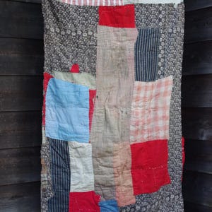 以下が含まれることがあります： さまざまな生地の柄と色を使ったパッチワークキルト。赤、青、白のパッチ、花柄、ストライプ柄が特徴です。キルトは木の構造に吊るされています。