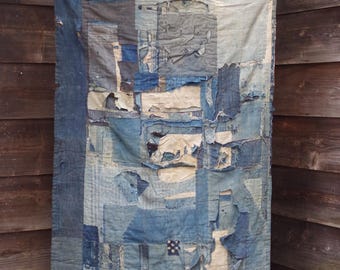 May include: A patchwork textile in shades of blue, white, and grey hangs against a wooden wall. The textile is made of various fabric pieces, some torn and distressed, creating a textured, vintage aesthetic. The piece is suspended from a thin, wooden rod.