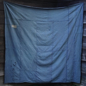 以下が含まれることがあります： 青と白のストライプの織物。キルトまたは壁掛けの可能性があり、風化した木の壁に展示されています。織物はパッチワークのデザインで、ステッチと小さなラベルが見えます。左下隅には日本語の文字が見えます。