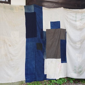 Könnte beinhalten: Textilkunstwerk aus verschiedenen Stoffflicken in Weiß, Marineblau und Grau. Das Patchwork-Design hängt vor einer Holzstruktur und zeigt eine Mischung aus Texturen und Farben. Das Stück ist wahrscheinlich ein Quilt oder eine Wandbehänge.