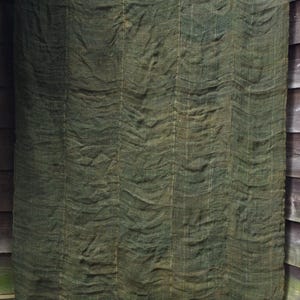 May include: A large, rectangular piece of dark olive green burlap fabric with a coarse, woven texture. The material features vertical seams and a series of horizontal folds or ripples throughout, hanging from a thin, light-colored wooden rod.
