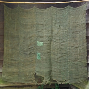 May include: A large, rectangular piece of olive green burlap fabric hanging from a horizontal bamboo pole. The coarse, woven material features vertical seams, two small rectangular green patches, and frayed, worn edges with holes.