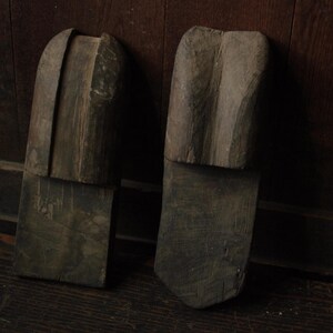 以下が含まれることがあります： 2つの風化した木製の靴型で、それぞれ2つの部分から構成されています。上部は湾曲して丸みを帯びており、下部は長方形です。木材は暗く、古びた外観をしており、アンティークまたはヴィンテージのアイテムを示唆しています。