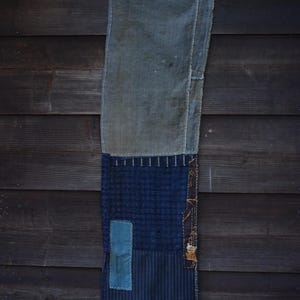 May include: A rectangular textile piece featuring a patchwork design of various blue fabrics. The top section is a faded grey-blue, while the lower portion consists of dark indigo grid patterns, vertical pinstripes, and a small solid light blue patch.