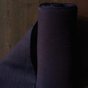 Puede incluir: Un rollo de tela de color morado oscuro con una textura acanalada vertical visible. La tela está enrollada firmemente en forma cilíndrica, con una parte del material desenrollada que se extiende hacia la izquierda, mostrando el color oscuro.
