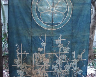 May include: A large, rectangular indigo-dyed textile with a floral emblem at the top and a grape vine design below. The design is in a lighter shade of beige. The textile is hanging outdoors, with trees and snow in the background.