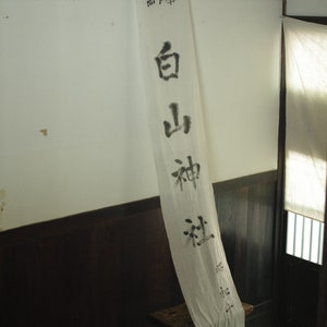 以下が含まれることがあります： 壁に掛けられた白い布の垂れ幕に、黒い墨で書かれた「錦  白山神社」の文字。これは「錦白山神社」と読みます。