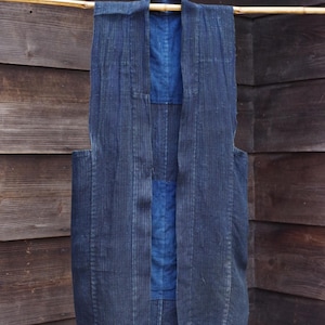 Puede incluir: Un chaleco azul índigo oscuro con un diseño de patchwork, colgado de una vara de bambú sobre un fondo de madera. El chaleco presenta un patrón de rayas verticales y un panel azul más claro contrastante.